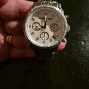 Silverfärgad klocka från Michael Kors - Stilren silverfärgad klocka från Michael Kors med vit urtavla och tre mindre urtavlor för extra funktioner. Klockan har en klassisk design med datumvisning och detaljerade visare. Perfekt för den som vill ha en elegant accessoar.