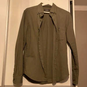 Olivgrön skjorta  - Snygg olivgrön skjorta, med klassisk krage och knappar framtill. Skjortan har en bröstficka med knapp och långa ärmar. Perfekt för en stilren look. Skick 8/10 använd sparsamt. Perfekt till sommaren med ett par linnebyxor 