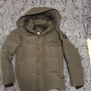 Grå dunjacka från Canada Goose - Säljer en grå dunjacka från Canada Goose med pälskantad huva. Jackan har en broderad logotyp på ärmen och stängs med dragkedja och knappar. Perfekt för kalla vinterdagar. Inga defekter. Är ute efter en snabb affär, där av priset! 