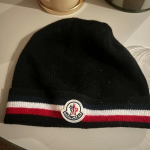 Svart mössa från Moncler - Snygg svart mössa från Moncler med broderad logga framtill. Mössan har en randig detalj i rött, vitt och blått runt kanten. Perfekt för kyliga dagar.