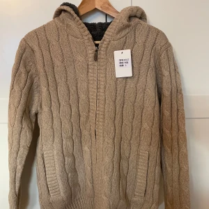 Beige stickad kofta med dragkedja - Mysig beige stickad kofta med dragkedja och huva från Maves. Tröjan har ett kabelstickat mönster och är fodrad med mjukt material för extra värme. Perfekt för kyliga dagar. Liten i storleken L men snarare som en M