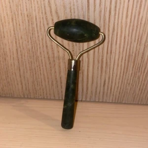 Grön ansiktsroller - Säljer en grön ansiktsroller i jade med ett handtag och rullhuvud i sten. Perfekt för att massera ansiktet och ge en svalkande känsla. Den har en elegant design med guldfärgad metall.