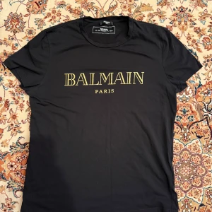 Svart t-shirt från Balmain Paris - Snygg svart t-shirt från Balmain med guldtryck av märkesnamnet på framsidan. Perfekt för en stilren och lyxig look. Kortärmad och tillverkad i mjukt material för bekväm passform. Storlek XL, passar L. Köpt på NK och använd ett fåtal gånger. Kvittot finns givetvis. 