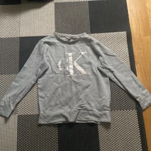 Grå tröja från Calvin Klein Jeans - Säljer en stilren grå tröja från Calvin Klein Jeans med det ikoniska CK-logotypen i vitt på framsidan. Tröjan har långa ärmar och en rund halsringning. Perfekt för en avslappnad look. Har lite avfärgning vid kragen.