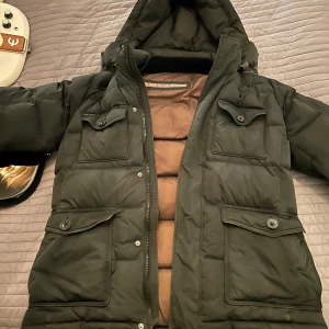 Svart dunjacka från Hetregó - Säljer en svart dunjacka från Hetregó med huva och flera praktiska fickor med knappar. Jackan har en dragkedja framtill och är perfekt för kalla dagar. Den är quiltad för extra värme och komfort. Passar S & M