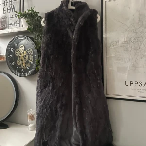Mörkgrå pälskappa - Säljer en stilren lång mörkgrå pälsväst som är perfekt för kyliga dagar. Västen har en mjuk och fluffig textur som ger en lyxig känsla. Den är ärmlös och har en klassisk design som passar till många olika outfits. Står inte storlek på men sitter som en S/M