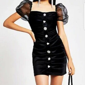 Svart sammet klänning - En svart klänning av materialet sammet. Klänningen har silverknappar framtill i olika former. Den har även volanger vid ärmarna. Pris kan diskuteras