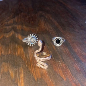 Tre unika ringar i olika stilar - Säljer tre unika ringar: en guldormring med detaljerad textur, en silverring med en svart oval sten omgiven av små stenar, och en silverring med ett solmotiv. Perfekta för att ge din stil en personlig touch.  20kr st.