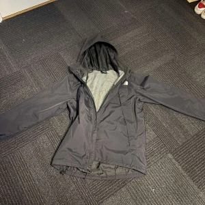 Svart vindjacka från The North Face - Säljer en svart vindjacka från The North Face med huva och dragkedja. Jackan har justerbara snören vid huvan och är perfekt för blåsiga dagar. Den är stilren och funktionell med The North Face-logga på bröstet. Nypris 1400