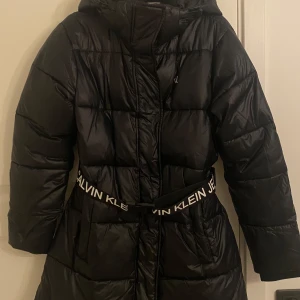 Svart dunjacka från Calvin Klein - Snygg svart dunjacka från Calvin Klein med huva och dragkedja. Jackan har ett bälte med logotypen 'Calvin Klein Jeans' som ger en stilren look. Perfekt för kyliga dagar. Jag har använt jackan bara 1 vinter, därav säljs den. Pris går och diskutera. (OBS! Senaste bilden är liknande variant av jackan nyare modell)
