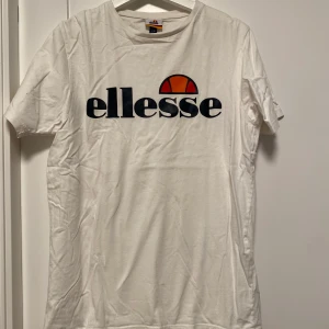 Vit t-shirt från Ellesse - Säljer en vit t-shirt från Ellesse med deras ikoniska logga i svart och orange på bröstet. T-shirten är kortärmad och tillverkad i bomull, perfekt för en avslappnad stil.