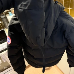 Canada Goose jacka  - Säljer en svart dunjacka från Canada Goose med huva och dragkedja. Jackan har en broderad logotyp på ärmen och är perfekt för kalla vinterdagar. Säljer en Canada Goose-jacka i storlek M. Färg: svart. Jackan är i nyskick och aldrig använd eftersom den är för liten för mig. Originalprodukt och mycket varm – perfekt för vintern! Priset kan diskuteras vid snabb affär.”