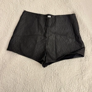 Skinnshorts - Snygga svarta skinnshorts med ett subtilt mönster och dragkedja i sidan. Från BikBok i storlek L men liten i storleken. 