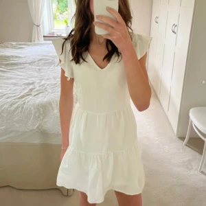 Vit klänning med volangdetaljer - Söt vit klänning med volangärmar och v-ringning. Den har en luftig passform och är perfekt för varma dagar. Klänningen har en knytning i midjan för en justerbar passform.