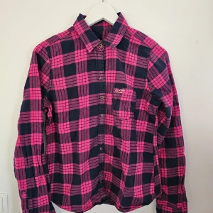 Rutig skjorta från Hollister - Snygg rutig skjorta från Hollister i svart och rosa. Skjortan har långa ärmar och knappar framtill. Perfekt för en avslappnad stil med en bröstficka som har en liten logga.