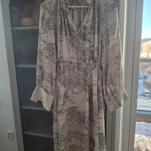 Beige mönstrad klänning från H&M - Säljer en elegant beige klänning från H&M med ett unikt svart mönster. Klänningen har långa ärmar och ett knytband i midjan för en smickrande passform. Perfekt för både vardag och speciella tillfällen.