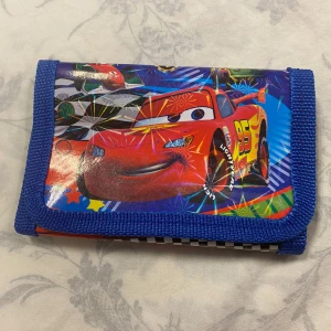 Cars Barn plånbok - Säljer en färgglad plånbok med motiv av en röd racerbil och nummer 95. Plånboken har en blå och röd kant och är perfekt för barn som älskar bilar. Den är kompakt och lätt att bära med sig. Finns fler kvar , samma frakt, 55kr/st . Helt ny 