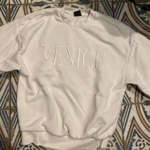 Vit sweatshirt med texten 'Venice' - Säljer en vit sweatshirt med texten 'Venice' broderad på framsidan. Tröjan har en rund hals och långa ärmar, perfekt för en avslappnad stil. Passar bra till både jeans och joggers.