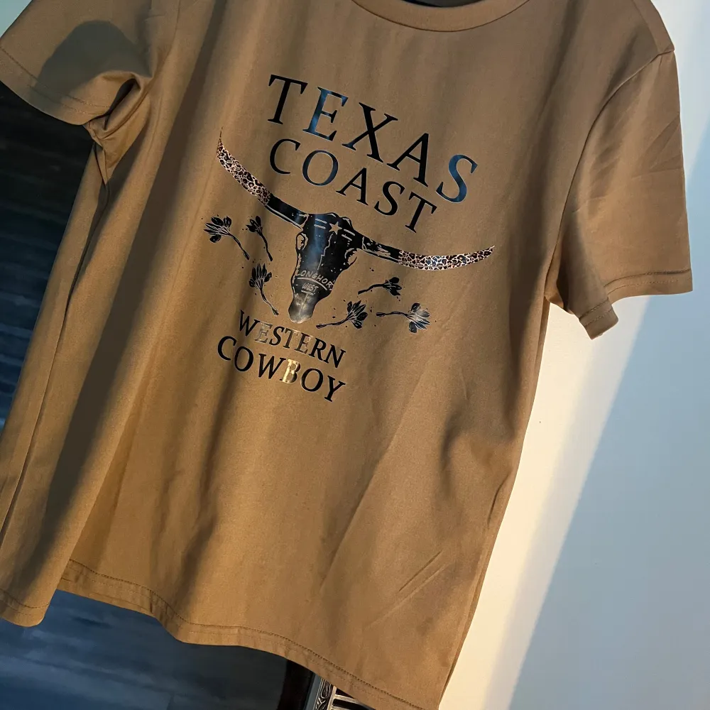 Säljer en beige t-shirt med tryck 'Texas Coast Western Cowboy'. Den har korta ärmar och är perfekt för en avslappnad stil. Materialet känns mjukt och bekvämt.. T-shirts.