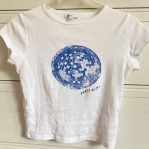 Vit t-shirt med blått discobolltryck från Subdued - Säljer en supercool vit t-shirt från Subdued i storlek S. Den har ett blått discobolltryck med texten 'Party Queen' på framsidan. Perfekt för en festlig look! T-shirten är kortärmad och gjord i mjukt bomullsmaterial. Den är i bra skick med bara ett litet märke på ärmen. Passar perfekt till både jeans och kjol!