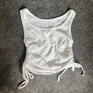 Vit ribbad linnetopp - Säljer en superfin vit ribbad linnetopp med knytning på sidorna. Perfekt för sommardagar eller att ha under en kavaj för en chic look. Toppen är ärmlös och har en tight passform som framhäver figuren. Ett måste i garderoben!