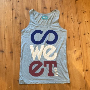 Grå linne från Sweets - Säljer ett ljusblått linne från Sweet SKTBS med ett stort tryck i blått, vitt och rött på framsidan. Linnet är ärmlöst och perfekt för sommardagar eller träning. Det är i storlek medium och i bra skick.