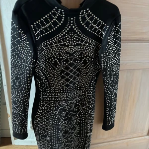 Svart klänning med paljetter - Säljer en supercool svart klänning med glittriga paljetter över hela framsidan. Den har en tight passform och långa ärmar, perfekt för en festkväll! Klänningen har en dragkedja framtill och är i nyskick.
