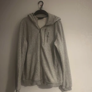 Grå hoodie från Sail Racing - Säljer en stilren grå hoodie från Sail Racing med dragkedja och en praktisk bröstficka. Tröjan har en bekväm passform och är perfekt för kyligare dagar. Den har en justerbar huva och ribbade muddar vid ärmslut och nederkant.