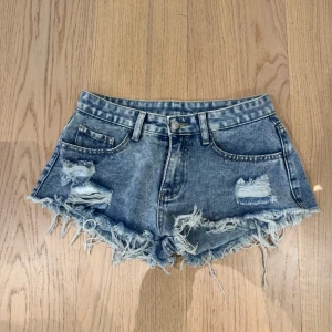 Blå lågmidjade jeansshorts med fransar - Snygga blå lågmidjade jeansshorts med slitna detaljer och fransar i nederkant. Perfekta för en avslappnad sommarlook. De har en klassisk femficksdesign och knappgylf.