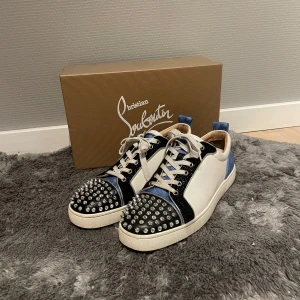 Christian Louboutin  - Snygga sneakers från Christian Louboutin i vitt, svart och blått. Skorna har en unik design med nitar på tån och en klassisk röd sula. Perfekt för den som vill sticka ut med stil. Skick 9/10, Hör gärna av dig vid minsta lilla fundering💯 