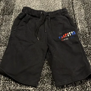 Svarta shorts från Trapstar - Snygga svarta shorts från Trapstar med färgglad logga på benet. De har en bekväm passform med snörning i midjan och är perfekta för en avslappnad stil. Materialet känns mjukt och skönt mot huden.