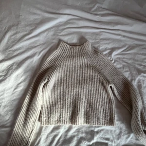 Beige stickad tröja från Monki - Säljer en mysig beige stickad tröja från Monki. Tröjan har en hög krage och långa ärmar, perfekt för kyliga dagar. Den är ribbad vilket ger en snygg textur och stil. Passar utmärkt till både jeans och kjol.