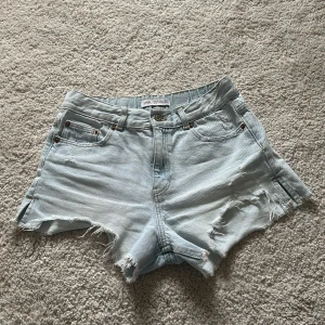 Ljusa jeansshorts från Zara - Säljer ett par ljusa jeansshorts från Zara med slitna detaljer. Shortsen har en klassisk femficksdesign och knappstängning fram. Perfekta för en avslappnad stil.
