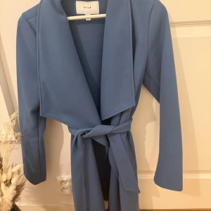 Blå kappa från Vila - Snygg blå kappa från Vila med knytband i midjan. Perfekt för att hålla stilen under kyligare dagar. Den har långa ärmar och en elegant krage som ger en sofistikerad look. Passar perfekt för både vardag och speciella tillfällen.
