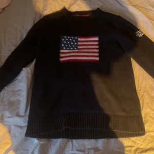 Mörkblå tröja med amerikansk flagga från Gant - Säljer en mörkblå tröja från Gant med en stor amerikansk flagga på framsidan. Tröjan är tillverkad i 100% bomull och har långa ärmar med ribbade muddar. Perfekt för en avslappnad stil.