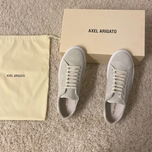 HELT NYA! (Oanvända) Beige sneakers från Axel Arigato - Helt nya och oanvända! Snygga beige sneakers från Axel Arigato i mocka med vit sula. Skorna har diskret snörning och märkets logga i guld på sidan. Perfekta för en stilren look. Nypris ca.2400kr, har box och bag till skorna.