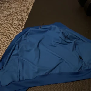 Blå hoodie från Hoodrich - Säljer en blå hoodie från Hoodrich med dragkedja och textdetaljer längs ärmarna. Perfekt för en avslappnad stil. Storlek M.