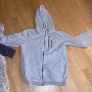 Sail racing hoodie - Säljer en grå hoodie med dragkedja och tryck på bröstet. Den har en praktisk huva och långa ärmar, perfekt för en avslappnad stil. Passar bra till både träning och vardag. 600 för både mökblåa o den här