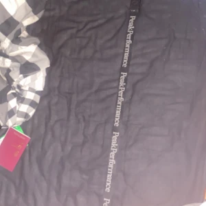 Svart bälte från Peak Performance - Snyggt svart bälte från Peak Performance med märkets logga tryckt längs hela bandet. Bältet har ett stilrent spänne med en diskret logotyp ingraverad. Perfekt för att ge en sportig touch till din outfit.