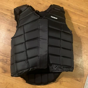 Svart skyddsväst från Jacson - Säljer en svart skyddsväst från Jacson. Västen har en quiltad design med justerbara remmar för optimal passform. Perfekt för ridning och andra aktiviteter där extra skydd behövs. Västen är både funktionell och stilren. Jag har aldrig haft chansen att använda den så den är helt ny. 