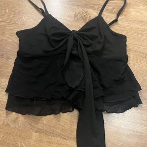Svart ärmlös topp med knyt - Snygg svart ärmlös topp med knyt framtill och volangdetaljer. Perfekt för en stilren look. Justerbara axelband för bästa passform.