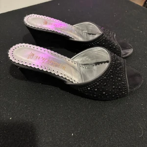 Svarta kilklackar - Eleganta svarta kilklackar från Whisper med öppen tå och dekorerade med små pärlor. Skorna har en bekväm kilklack och en glansig innersula i silver. Perfekta för en stilfull look.