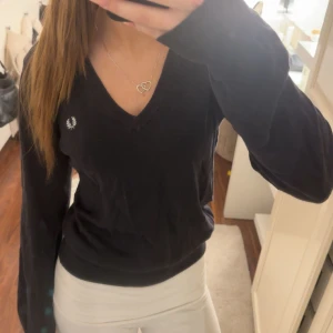 Svart tröja från Fred Perry - Säljer en snygg svart/mörkblå tröja från Fred Perry med v-ringning och logga på bröstet. Tröjan har långa ärmar och är perfekt för en avslappnad men snygg look. Passar bra till både jeans och kjol. Lappen i nacken sitter ganska löst☺️sitter som storlek Xs/s nypris runt 1300kr