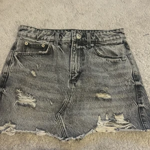 Grå jeanskjol från Zara - Snygg grå jeanskjol från Zara med slitna detaljer och rå kant. Perfekt för en trendig look. Kjolen har klassiska femficksdesign och knappgylf.