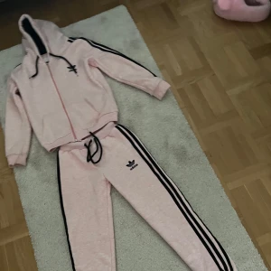 Rosa hoodie och byxor från Adidas - Säljer en snygg rosa hoodie och byxor från Adidas med svarta detaljer och dragkedja. Tröjan har långa ärmar och klassiska Adidas-ränder längs ärmarna, så även byxorna. Perfekt för en sportig look! 