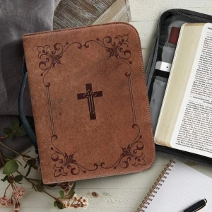 Brun bokväska med korsmotiv - Elegant brun bokväska med ett korsmotiv på framsidan. Väskan har flera fack för pennor, anteckningsblock och andra tillbehör. Perfekt för att bära med sig en bok eller bibel. Den har en praktisk bärrem och dekorativa mönster i hörnen.