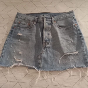 Jeanskjol från Levi's - Snygg jeanskjol från Levi's i ljusblå denim med slitna detaljer och fransig nederkant. Kjolen har knappar framtill och klassiska femficksdesign. 