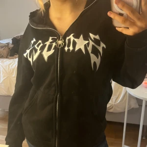 Svart hoodie med spindelmotiv - Säljer en svart hoodie med dragkedja och ett stort vitt spindelmotiv på ryggen. Framsidan har ett tryck i vitt med stjärnor och text. Huvan har vita detaljer som liknar ögon. Perfekt för en unik och cool stil!