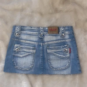 Blå jeanskjol från Gallop - Snygg blå jeanskjol från Gallop med slitna detaljer och dekorativa fickor både fram och bak. Kjolen har en knapp och dragkedja framtill samt bälteshällor för extra stil. Perfekt för en avslappnad look!