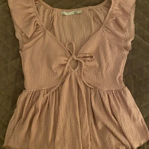 Ljusrosa topp med volangdetaljer - Säljer en söt ljusrosa topp med volangdetaljer och knytning framtill. Toppen är perfekt för varmare dagar och den är tillverkad i ett lätt och bekvämt material🥰 Bara använd en gång